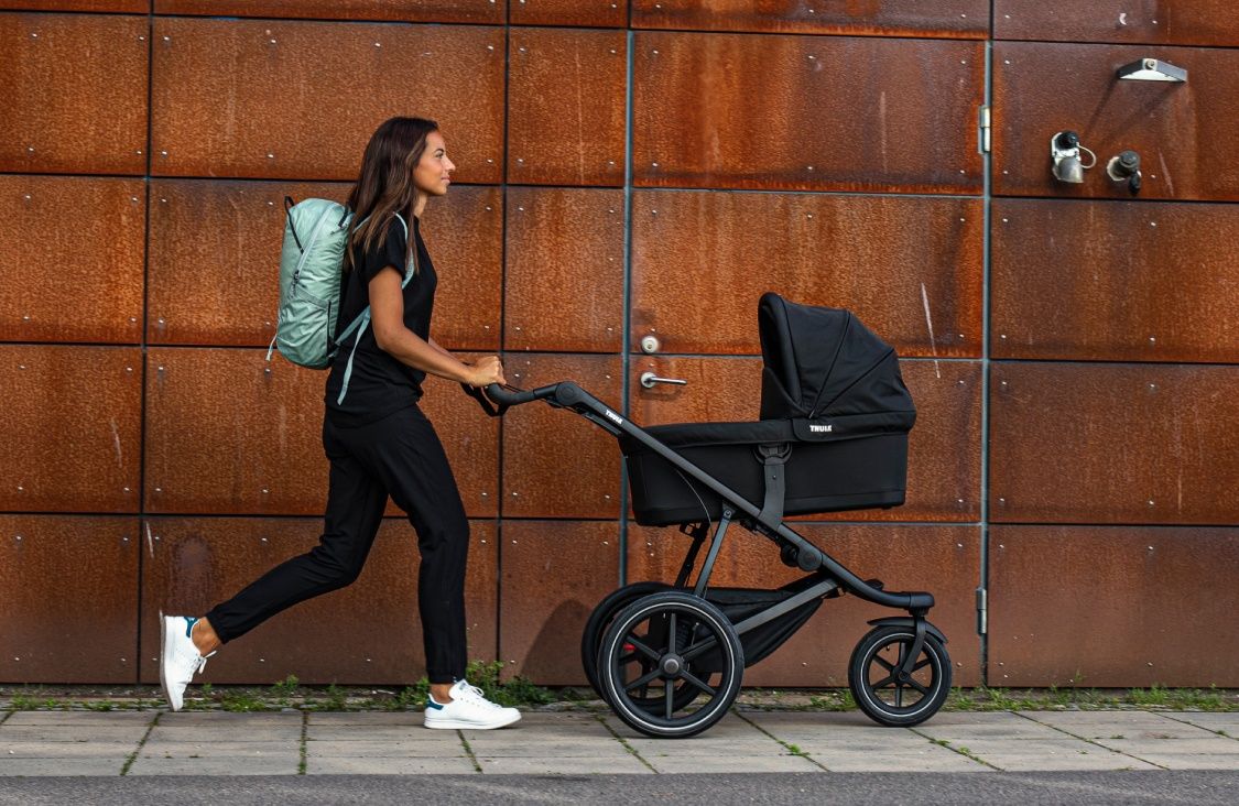 A woman walks down a city street doing a postpartum exercise of walking with a black bassinet stroller.