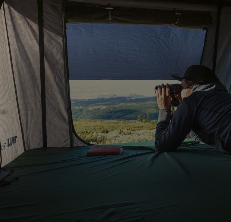 A man inside a Thule Tepui roof top tent looks out at the mountains with his binoculars.