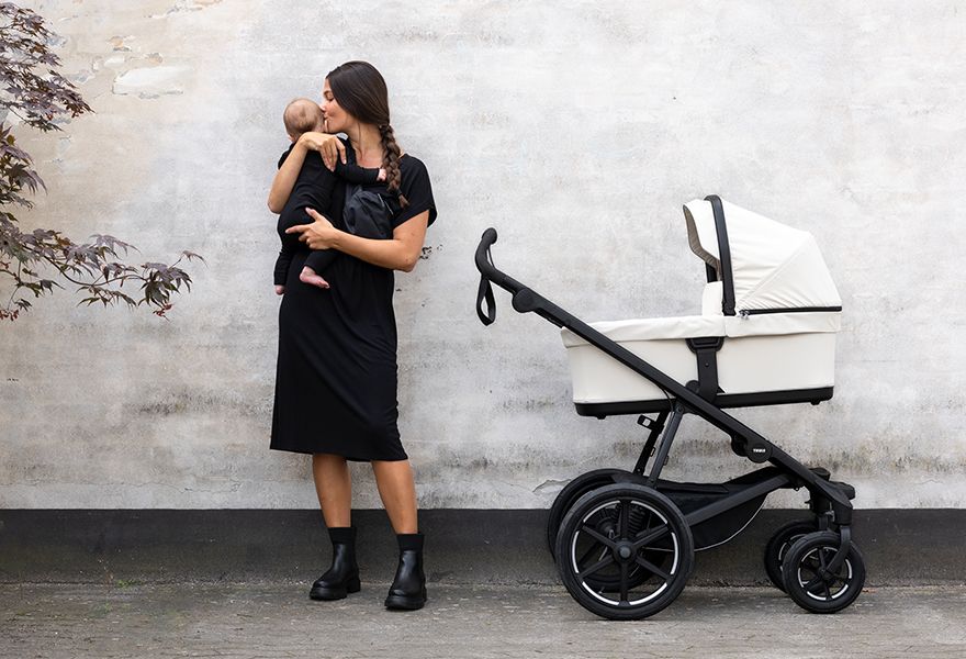 A woman is holding a baby next to the Thule Urban Glide 4-wheel stroller.