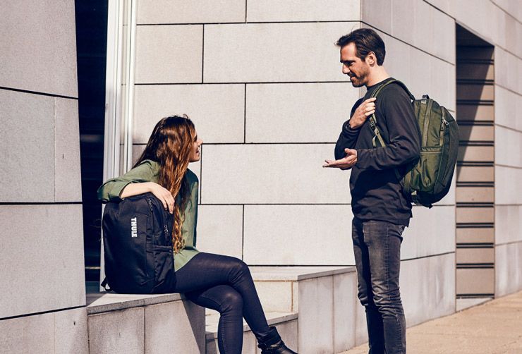 A woman sits on a ledge with a Thule backpack, while a man speaks to her also carrying a Thule backpack.