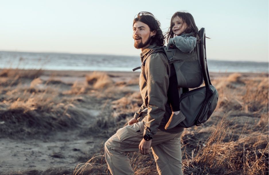A man walks by the beach with his child in a Thule Sapling child carrier backpack.