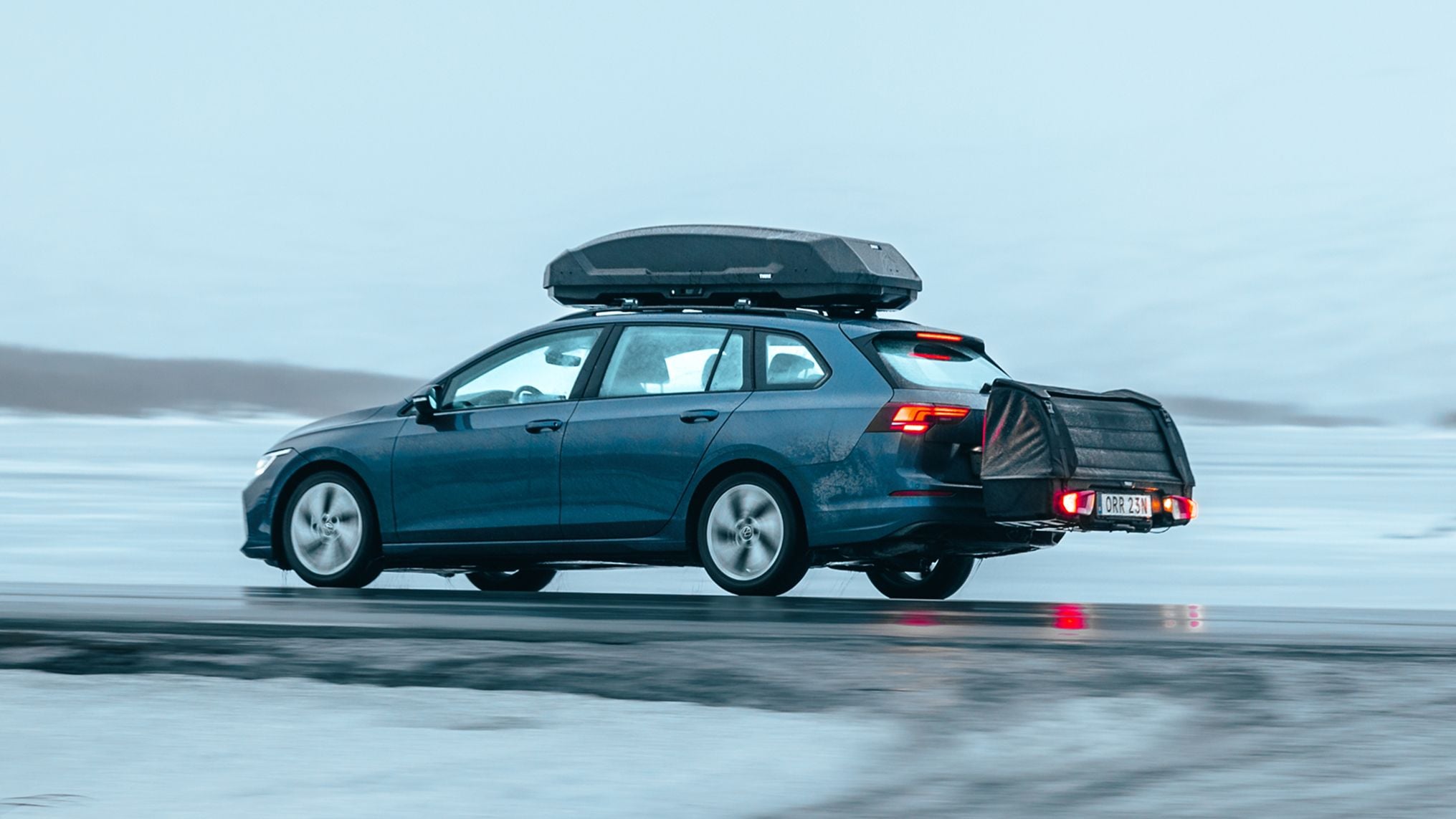 A blue car driving on a wet, icy road with a roof box and a rear‑mounted cargo carrier attached, set against a snowy landscape.