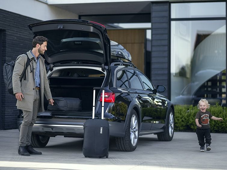 A man stands with his Thule carry on luggage beside and a parked car where a child is running.