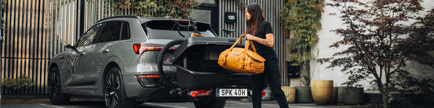 在都市场景中，一名女子将黄色旅行包放入灰色汽车上安装的 Thule Onto 2 后置载物箱中，还有一辆自行车。