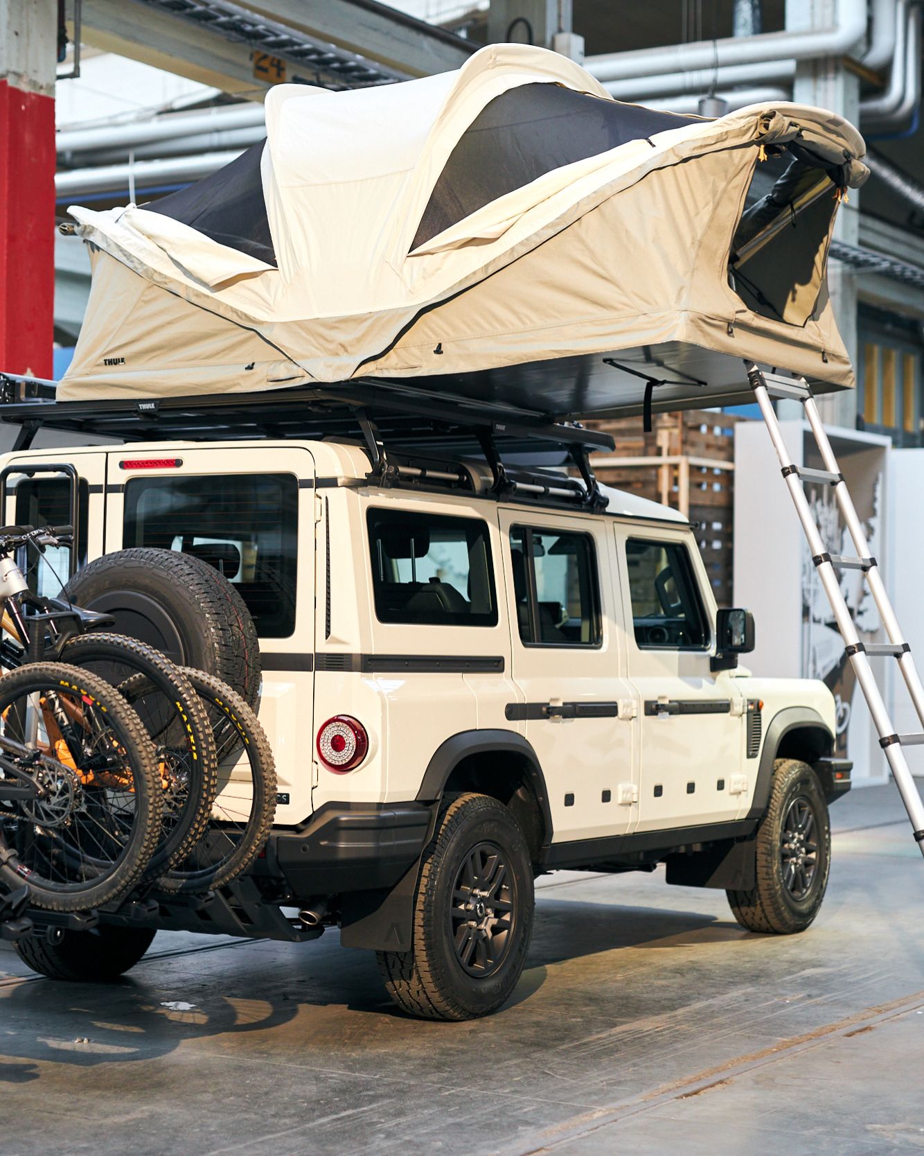 A white jeep parked in an exhibition hall with a an unfolded Thule Approach rooftop tent and three bicycles on the back.