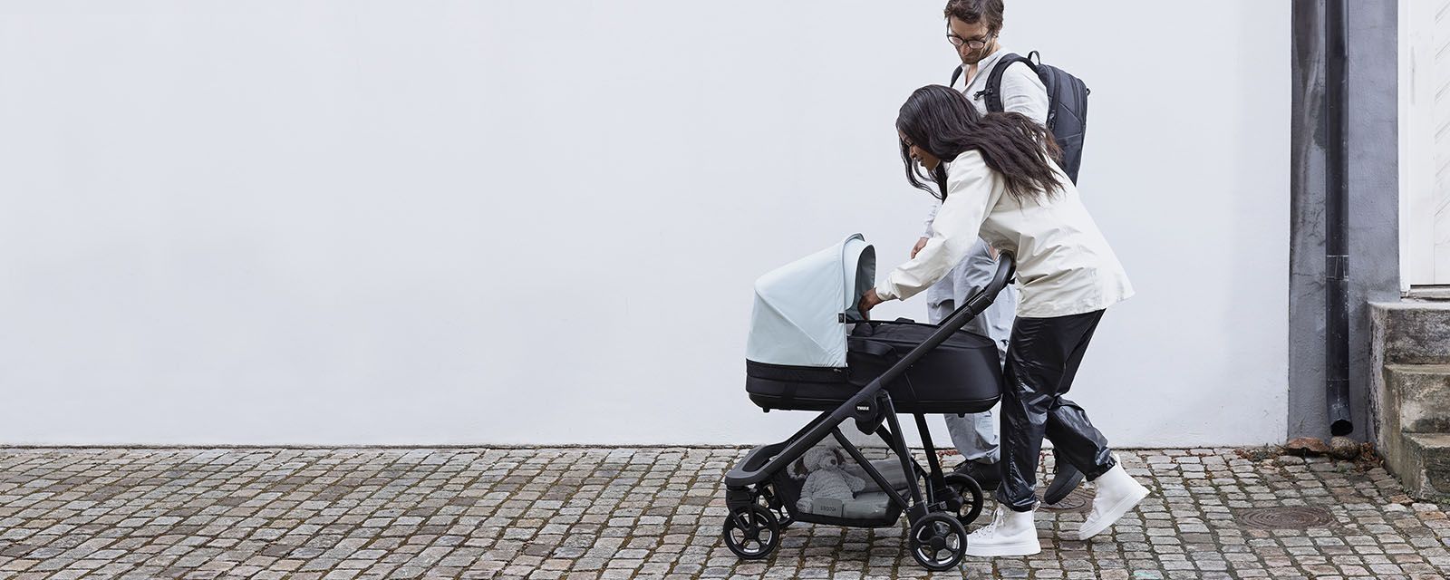 A couple walk down a cobbled street and look at their baby in a Thule Shine bassinet stroller.