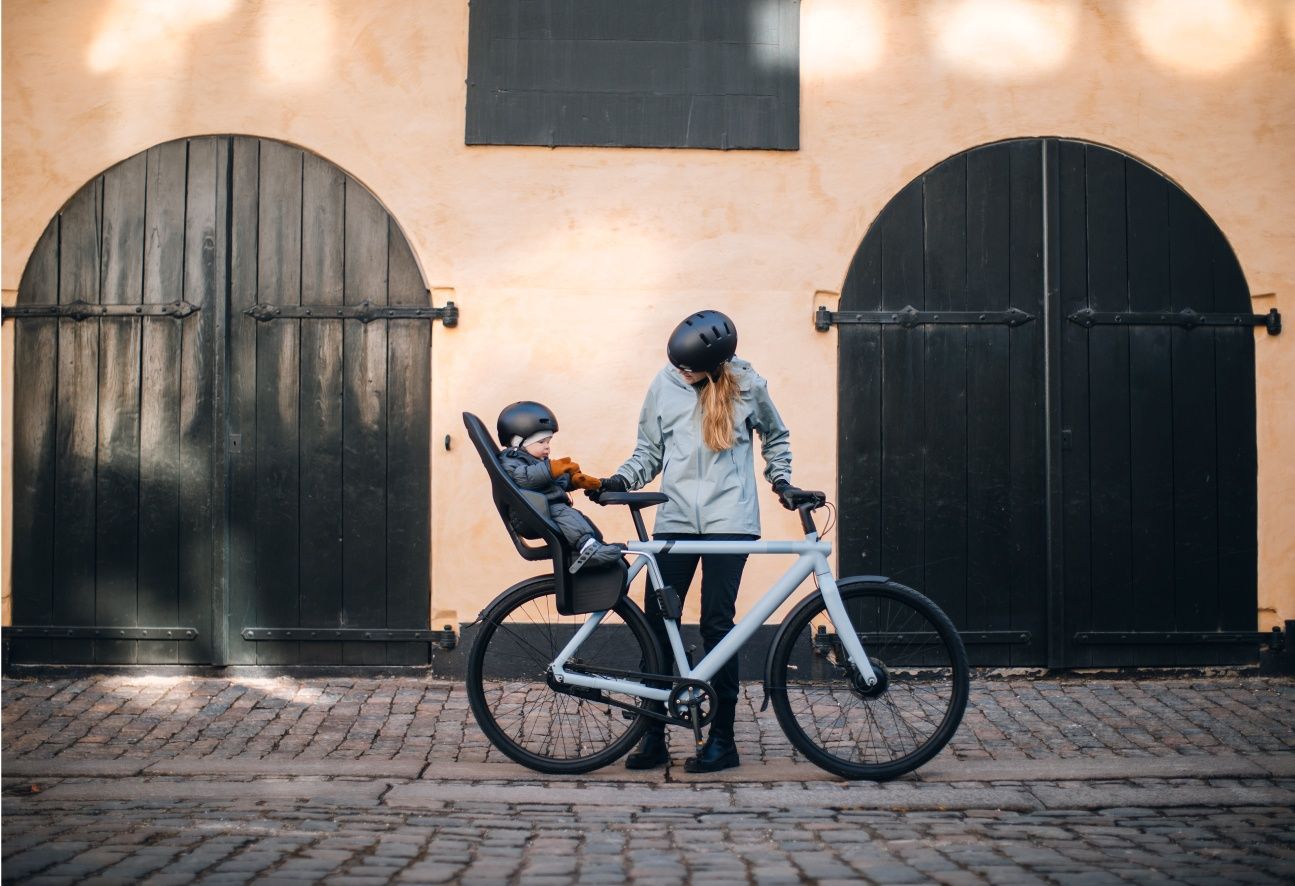 A child sits in a rear Thule child bike seat attached to a bike while a woman stands next to the bike.