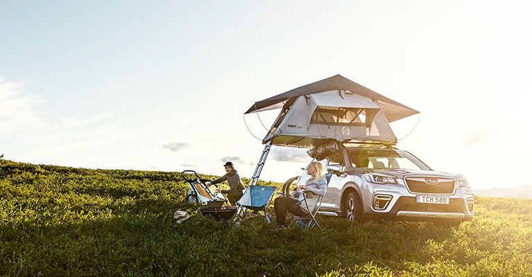 A family sits under a Thule rooftop tent that is parked in a field with the sun setting.