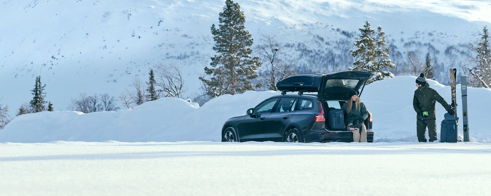 Two people stand by a car parked in the snow holding ski boot backpacks, and ski bags.