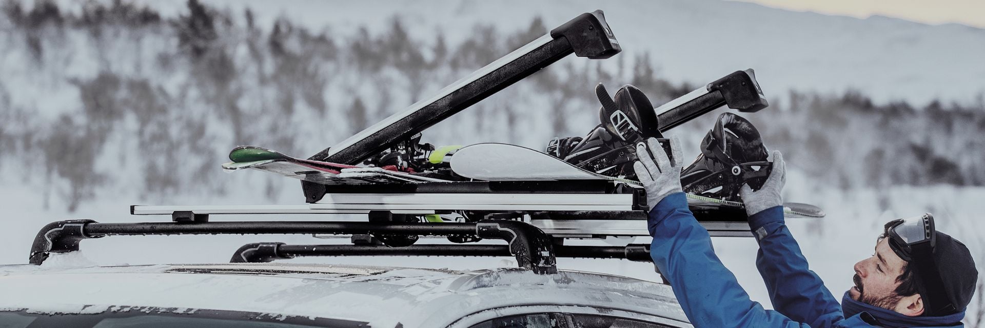 Person loading skis onto a Thule roof platform on a car in a snowy mountain landscape. 