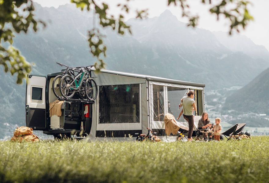 A motorhome is parked in a field with an awning tent and a Thule VeloTrack bike rack.