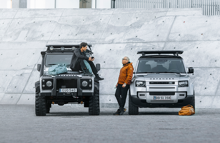 Two men stand next to vehicles with the Thule Caprock roof platforms chatting.