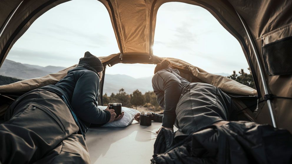 Two people sit in a soft shell roof top tent with panoramic skylights looking at the mountain view.
