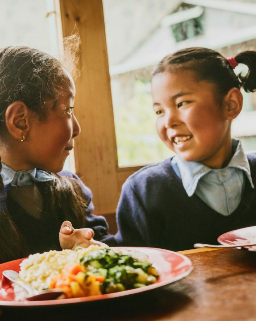 Two young girls smile at each other while eating a meal.