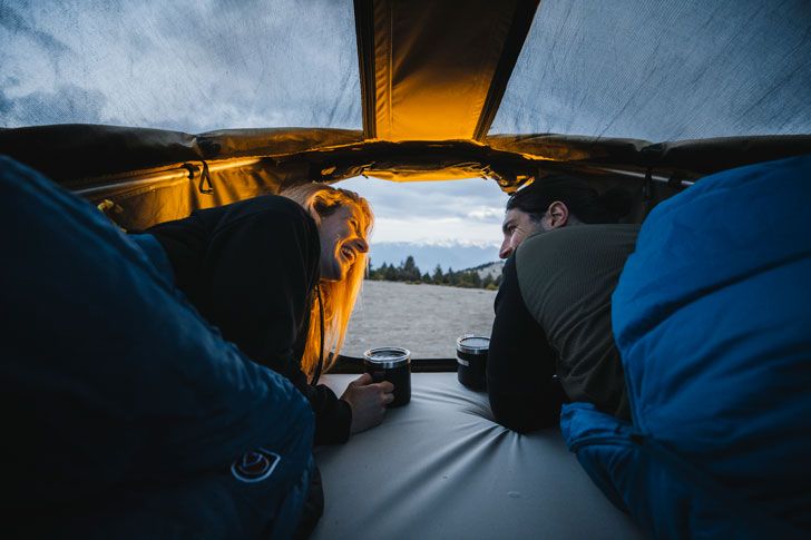Deux personnes allongées à l’intérieur d’une tente de toit avec des sacs de couchage boivent des boissons chaudes dans des tasses.