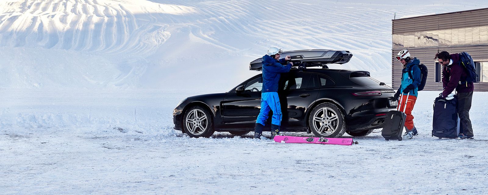 I snevejr ses to mennesker med skiudstyr. De er ved at laste deres ting ind i en bil og en tagboks.