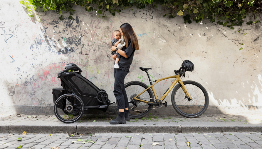 Person, der holder en baby ved siden af en parkeret cykel og en børnetrailer.