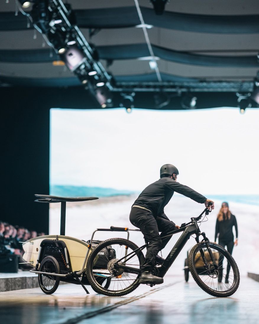 A person riding a bike with a cargo trailer on a runway in front of a large screen displaying a landscape.