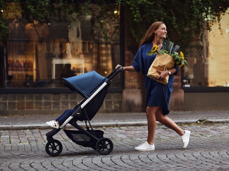 A woman wearing a blue dress holding a bag of groceries, pushes her compact stroller down a cobbled street.