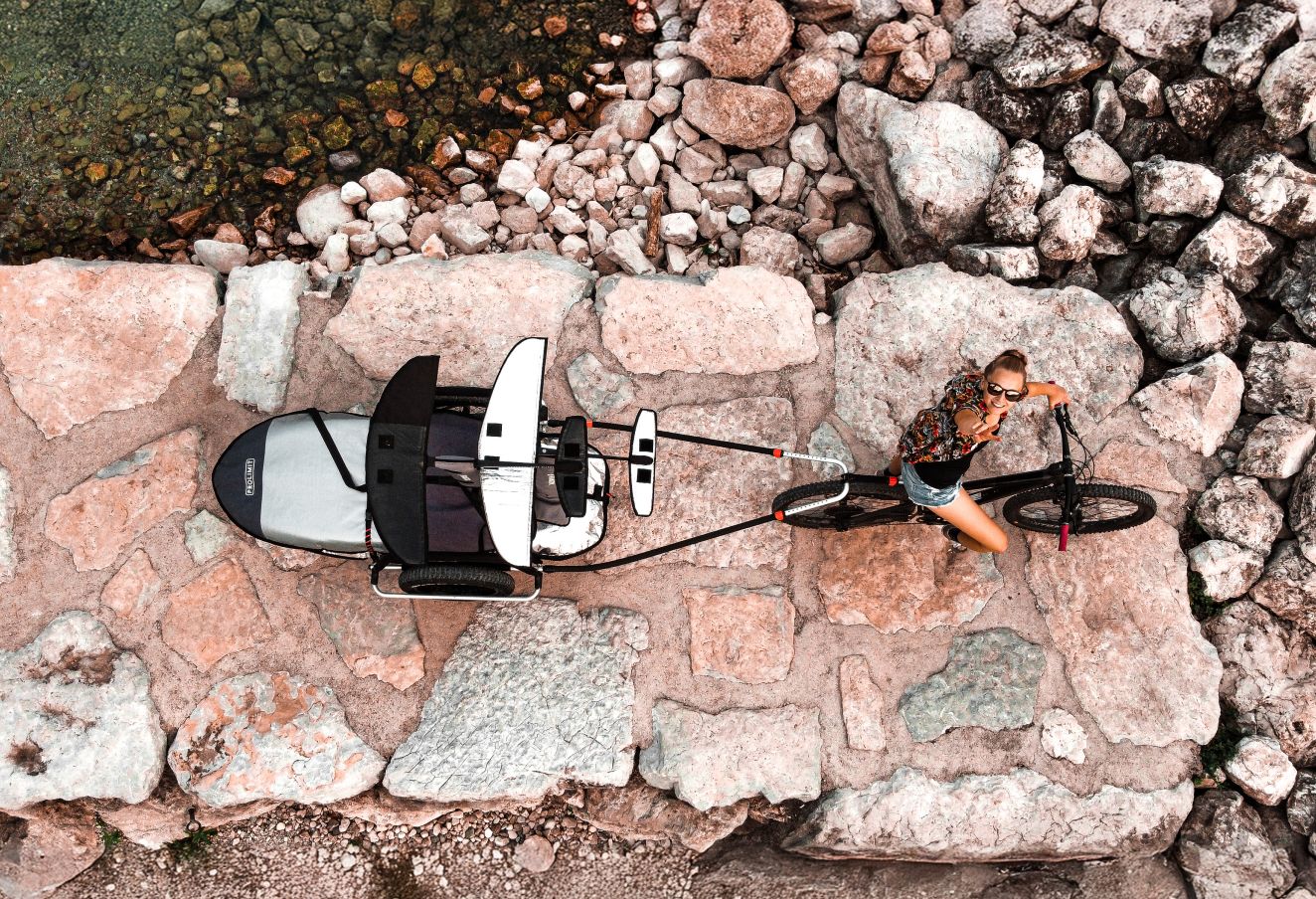 Ansicht von oben auf einen Reacha Sport Fahrradanhänger, der auf felsigem Gelände in der Nähe des Wassers geparkt steht und mit Paddel- und Surfbrettern beladen ist.