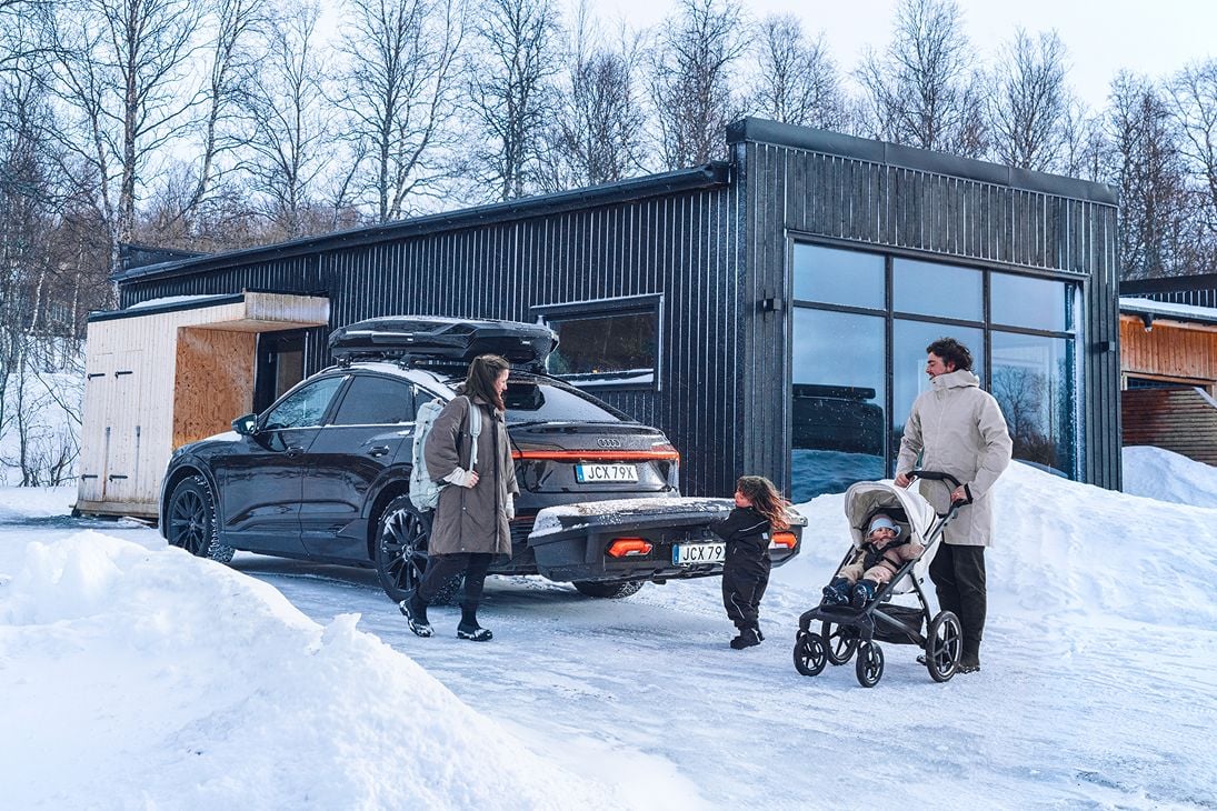 Vuxen som skjuter en barnvagn och ett barn som går mot en bil med en roodbox och en Thule Arcos bakmonterad lastbärare.