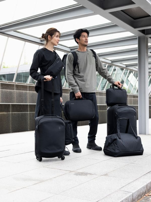 A stylish couple stands outside a modern transportation hub, each carrying and wheeling various pieces from the Thule Subterra 2 luggage collection.