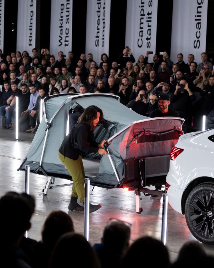 A woman unfolding a Thule Outset towbar mounted tent on a fashion runway with a crowd of people looking at her.