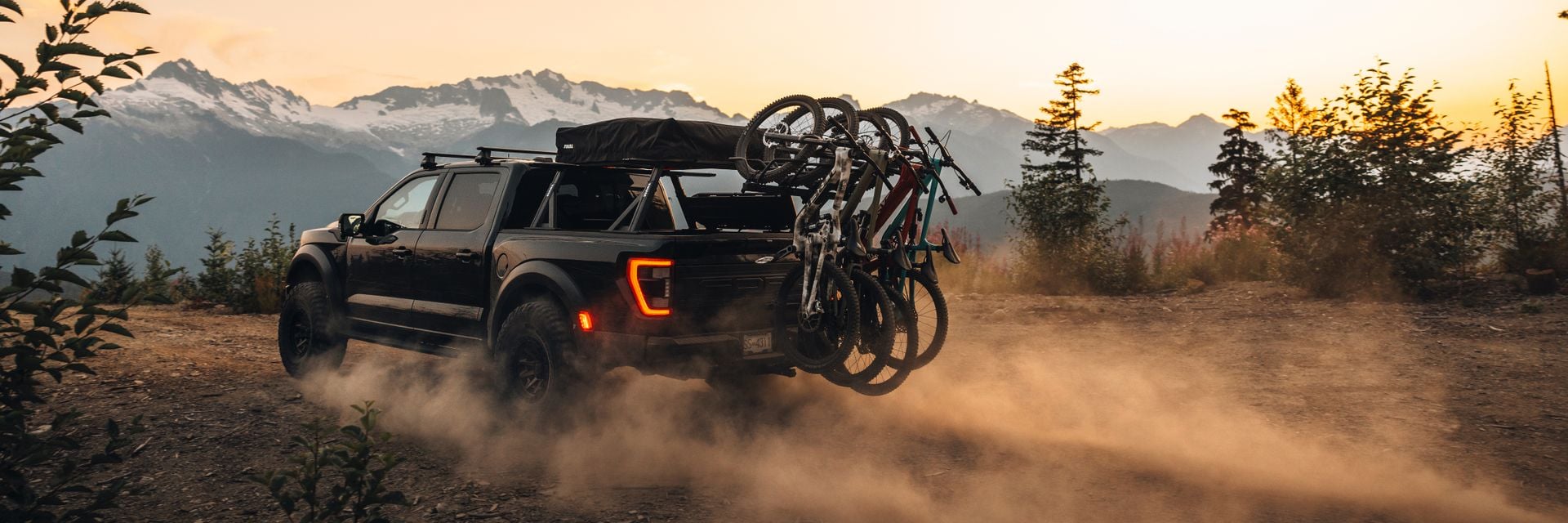 Camioneta conduciendo por una carretera montañosa polvorienta al atardecer con varias motos montadas en un portabicicletas Thule Revert, promocionando Bring Your Bike.
