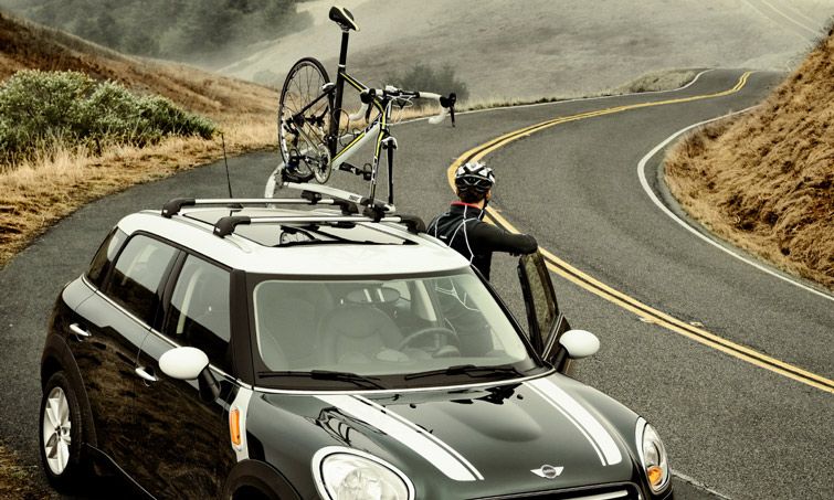 On a street a cyclist gets out of a car with Thule AeroBlade Edge roof racks and a fork mount bike rack attached.
