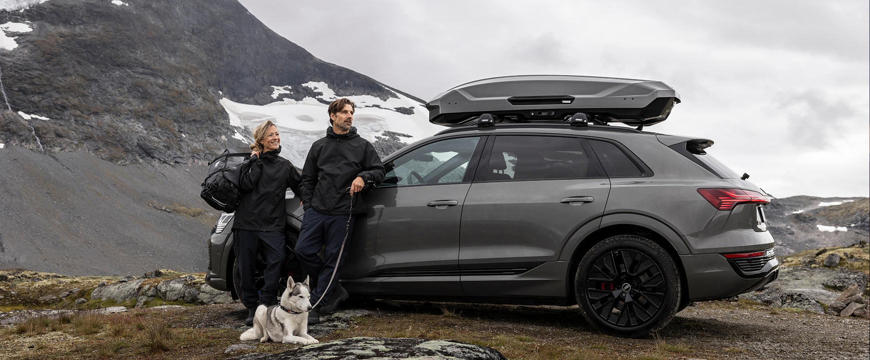 Um SUV escuro com caixa de teto está estacionado em uma encosta rochosa de montanha, enquanto duas pessoas estão ao lado do veículo com equipamentos externos e um cachorro sentado aos seus pés. Manchas de neve e montanhas íngremes cercam a cena sob um céu nublado.