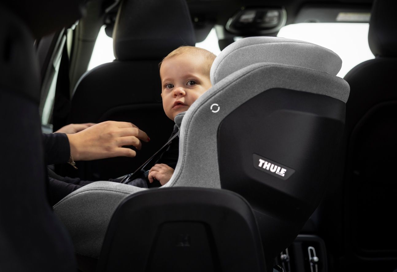 A young toddler sitting in a Thule Elm toddler car seat in light grey. A person's hands are near the toddler's straps.