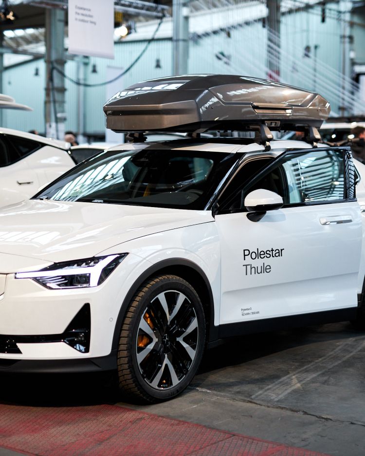A white Polestar 2 parked in a large exhibition hall with a Thule Motion 3 roof box in a dark grey color on top of it. There are other white cars parked behind and next to this one.