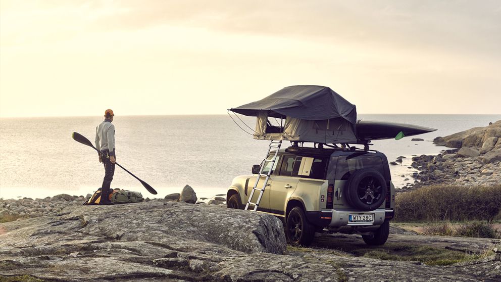 A man with a kayak paddle is standing next to his vehicle by the sea with a Thule rooftop tent  and kayak mounted to the roof.