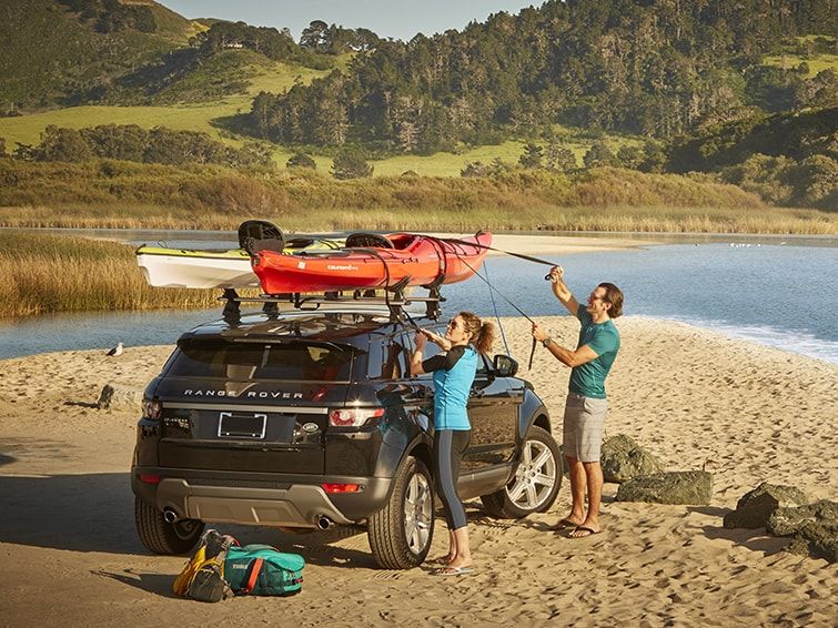 Next to a beach by a hill, a couple secure their kayak on a kayak roof rack on a car.