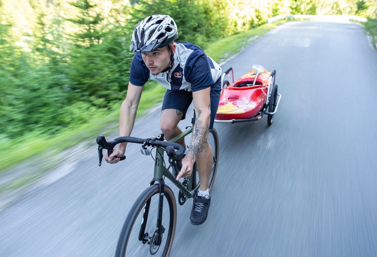 Un ciclista circulando por un camino en un bosque con un remolque para bicicleta Reacha Sport que lleva un kayak rojo, destacando su estabilidad en terrenos al aire libre.