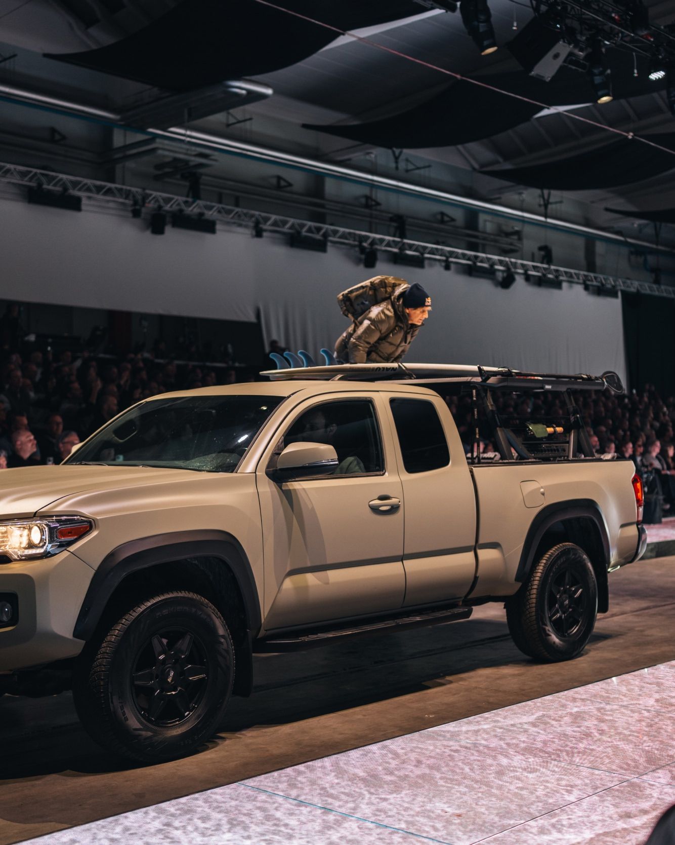 A pickup truck with mounted gear driving onto a runway during a show, with an audience watching.
