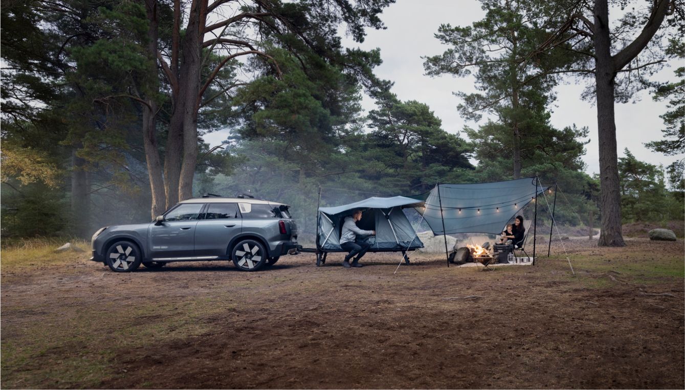 两人在森林中露营，有一顶 Thule Outset 帐篷与灰色 SUV 相连；一个人在帐篷里，另一个人坐在篝火旁的遮阳篷下。