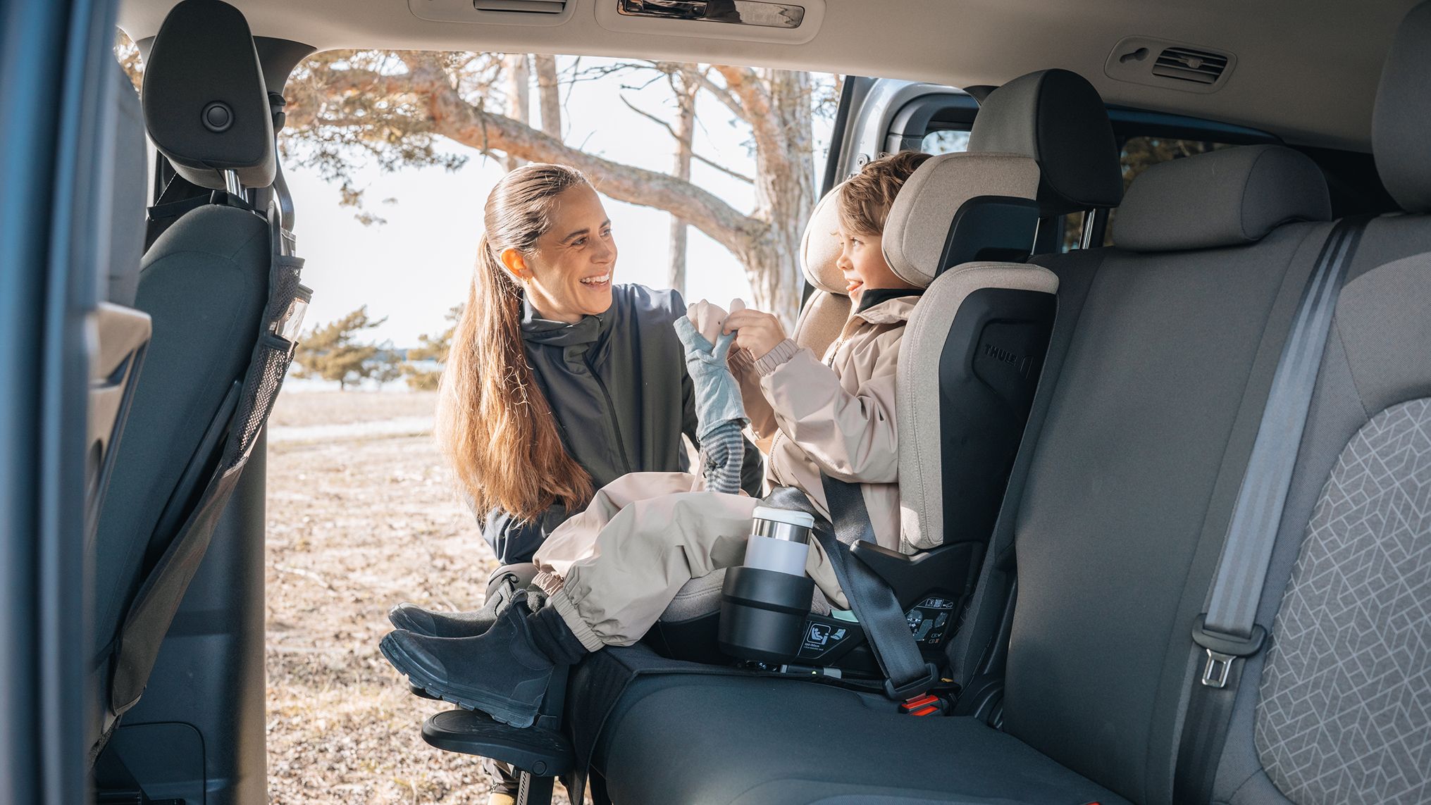 Anna Haag sorrindo para uma criança sentada em um assento infantil Thule no banco de trás de um carro, com a porta aberta para uma paisagem arborizada.