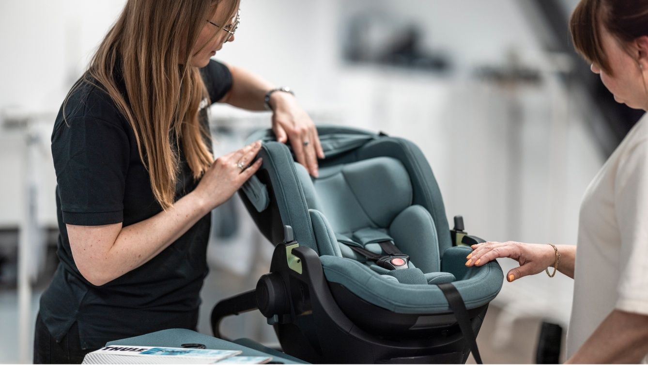 Due donne esaminano l'aderenza e le caratteristiche di un seggiolino per auto rivolto all'indietro in un ambiente interno controllato.