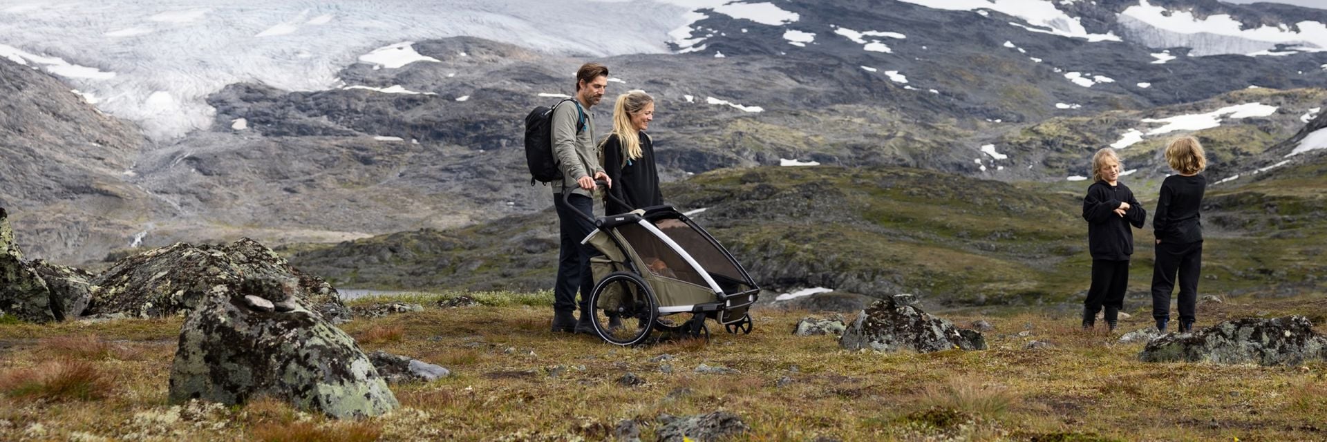Con un telón de fondo montañoso, dos adultos están junto a un carrito Thule con un bebé dentro mientras dos niños juegan cerca.
