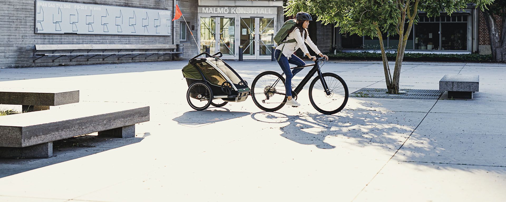 Une femme portant un sac à dos roule à vélo en ville avec une remorque Thule Chariot Cab.