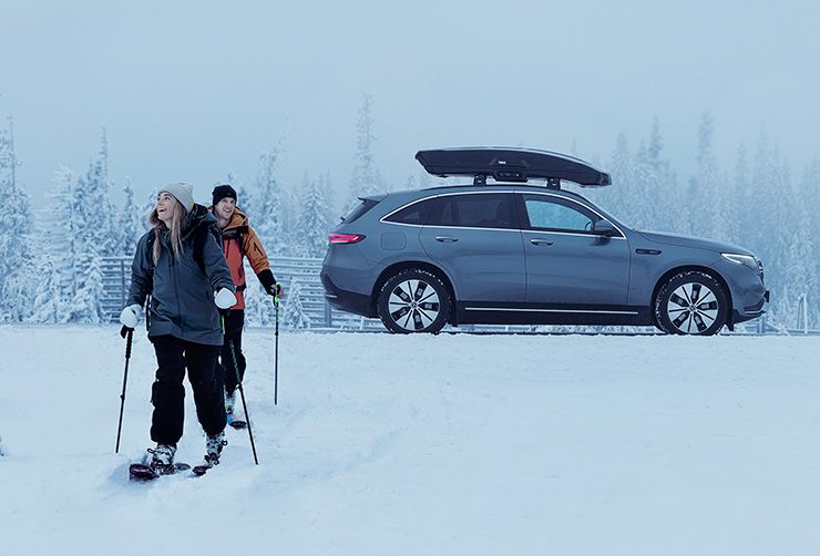 Deux skieurs s'en vont et on aperçoit derrière eux leur voiture électrique équipée d'un coffre de toit pour skis.
