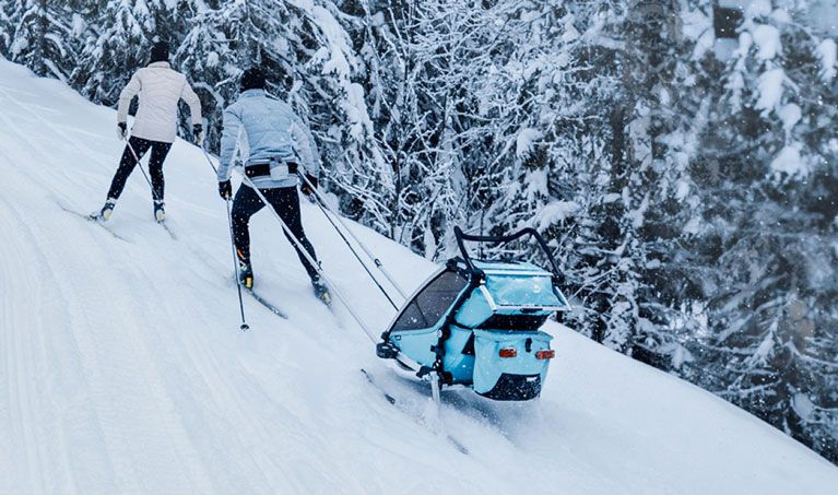 Two cross-country skiers ski up a hill with the multisport trailer Thule Chariot Cross.
