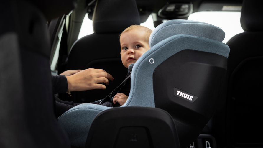 Noor väikelaps, kes istub sinises Thule Elm väikelapse turvatoolis. Väikelapse rihmade lähedal on inimese käed.