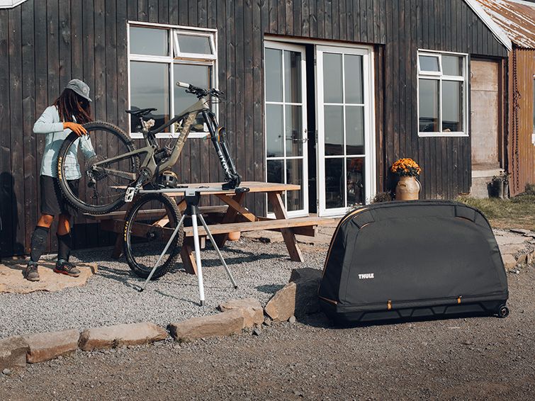 Next to a wood building a cyclist fixes her bike on a bike stand next to the Thule RoundTrip bike travel case.