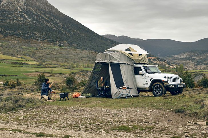 A jeep is parked in the mountains with a roof top tent and a woman sits under the annex while a man grills.