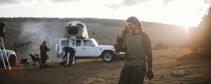 Un hombre pasea con una bebida caliente, al fondo hay personas que montan un campamento alrededor de una tienda para coche Thule Approach