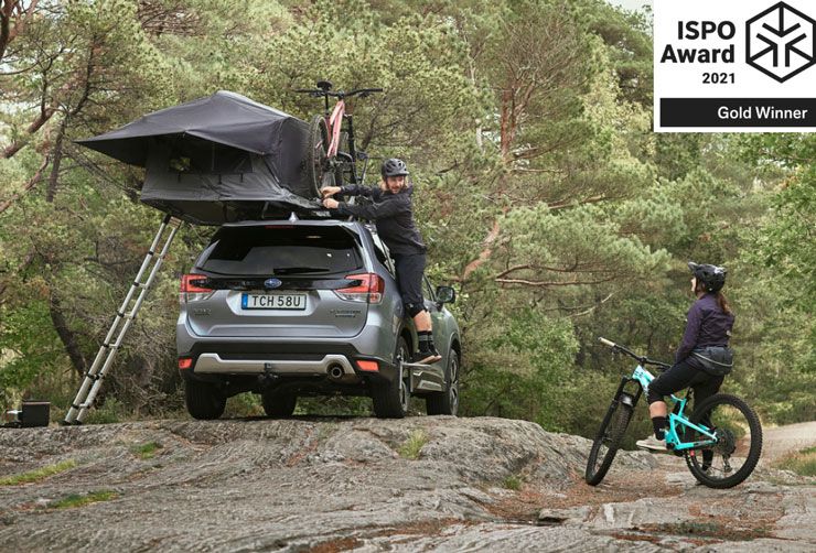 Man in a forest unloads bike from car roof where a Thule Tepui Foothill roof top tent is also mounted.