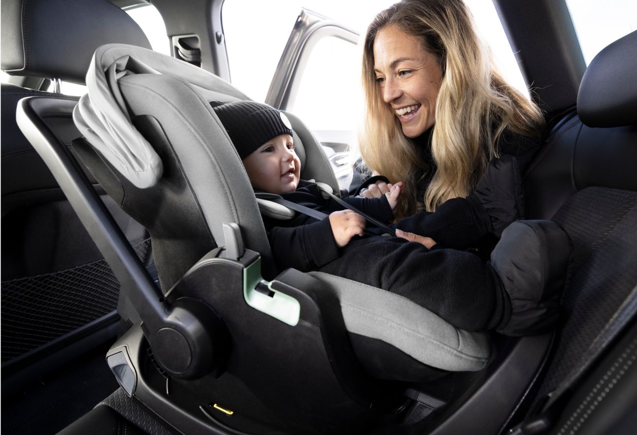 A blonde-haired woman leaning inside a car and smiling. In the car is a baby placed inside the Thule Maple infant car seat, facing rearwards.