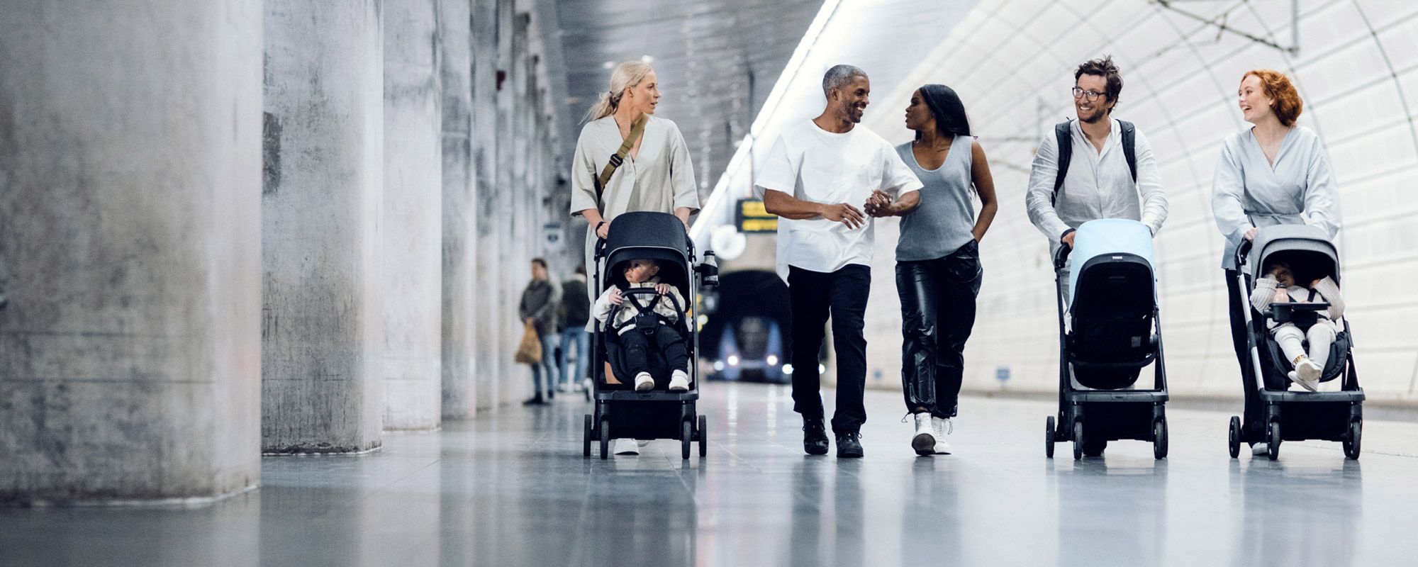 A group of parents at a train station push their children in Thule Shine strollers.